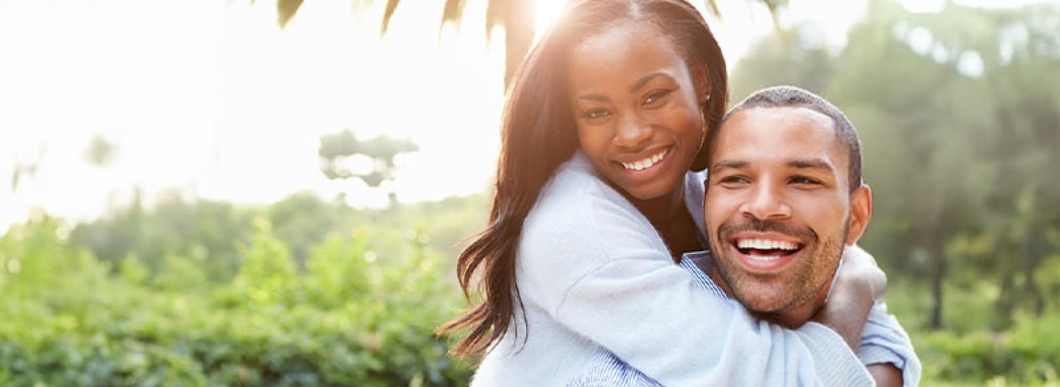 A happy man and woman outside in the sun. She has her arms wrapped around him.