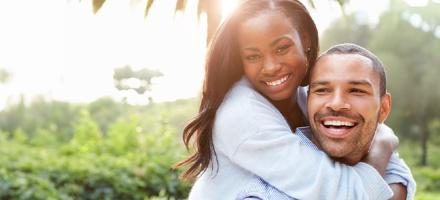 A happy man and woman outside in the sun. She has her arms wrapped around him.